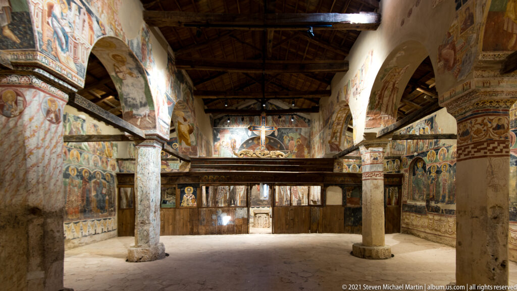Oblique nave interior with frescoed columns, arches, mural-covered walls, timber roof, and wooden sanctuary screen.