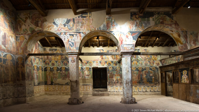 Three interior arches on painted columns facing mural-covered walls and a central dark doorway.