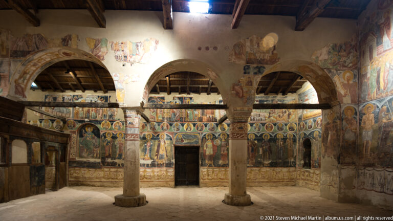 Frescoed nave arcade with two central columns, saintly mural registers, timber roof, and wooden screen at left.