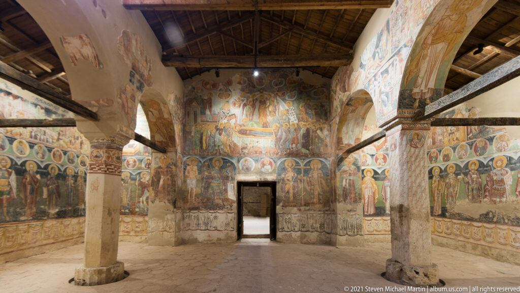 Symmetrical church interior with central doorway, painted arches, columns, rows of saints, and timber roof.