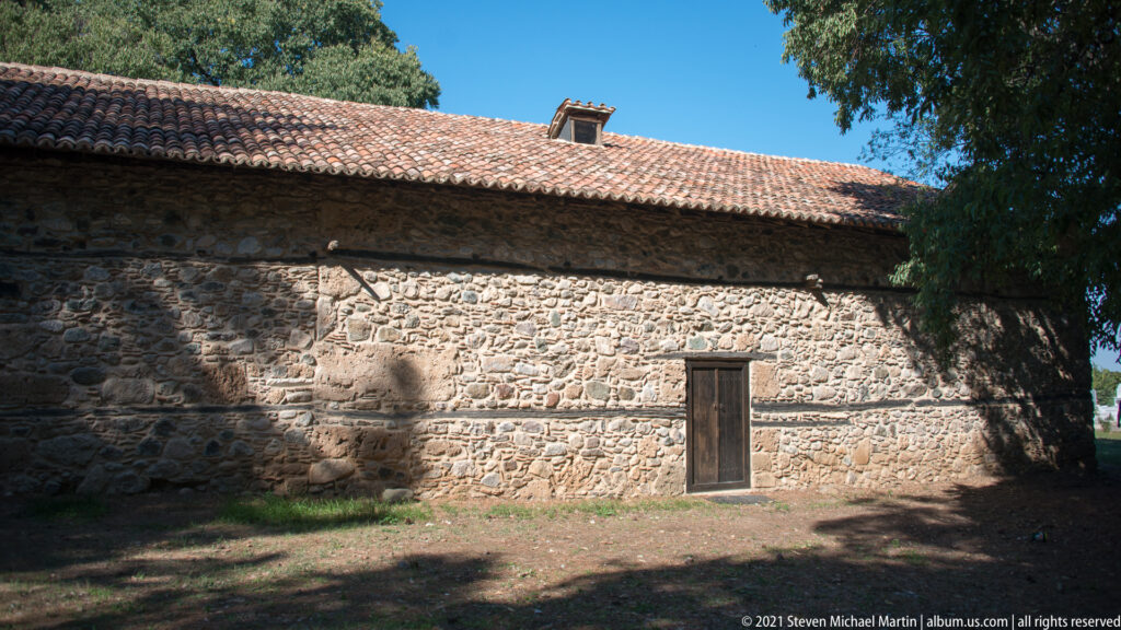 Long fieldstone side wall with a wooden door, low red-tiled roof, and deep tree shadows.