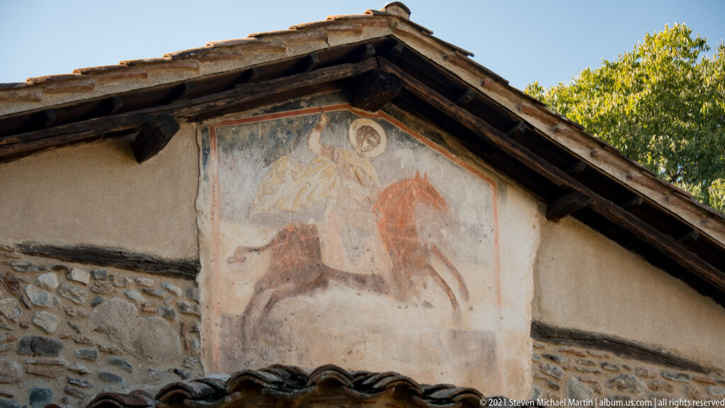 Faded exterior fresco of a haloed rider on a reddish horse beneath the projecting gable roof.