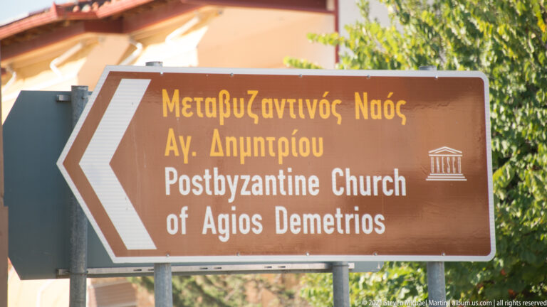 Brown bilingual heritage sign reading “Postbyzantine Church of Agios Demetrios” with a left-pointing arrow.