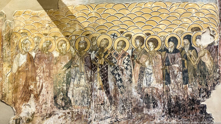 Large wall painting of many standing haloed saints arranged closely in rows against a gold-toned background.