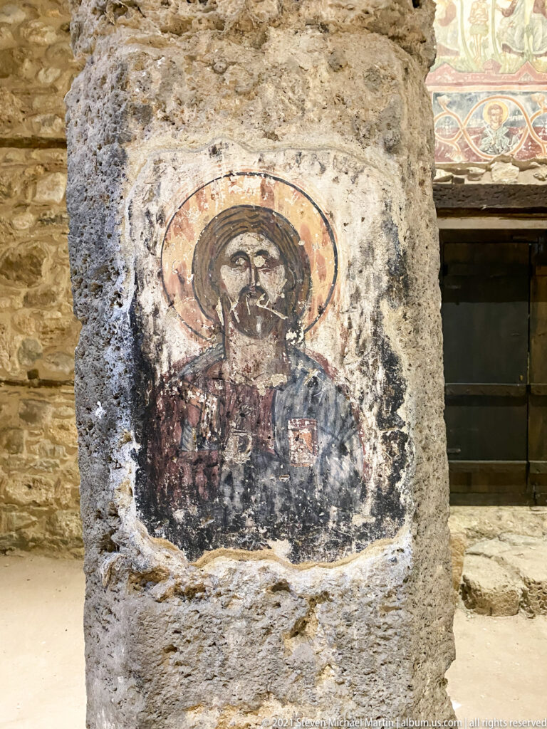 Half-length haloed saint painted on a rough pier, with one hand raised and the surrounding plaster heavily worn.