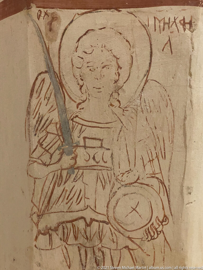Incised red-line drawing of a haloed winged figure holding a rectangular book, with scratched letters nearby.