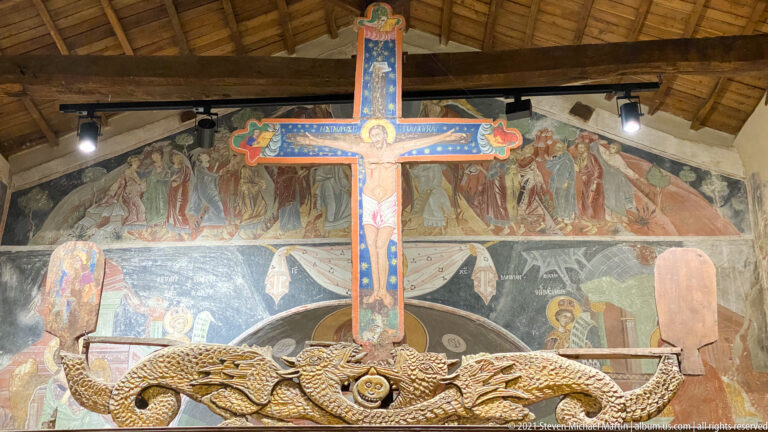 Painted Crucifix above carved furnishings with a large multi-register fresco covering the wall behind.