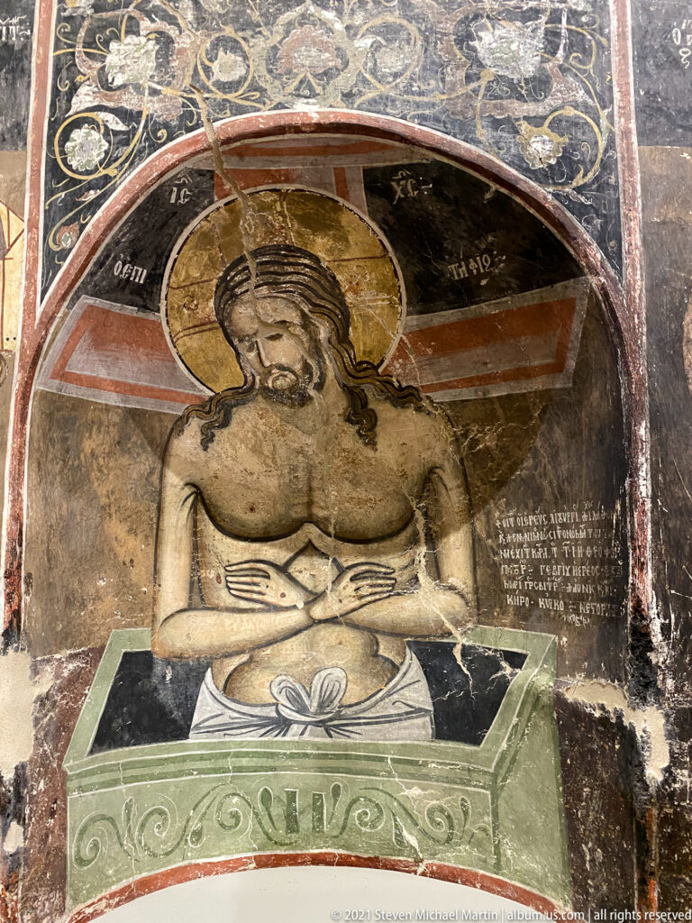 Half-length Christ with bowed head and crossed hands emerging from a painted sarcophagus beneath an arch.