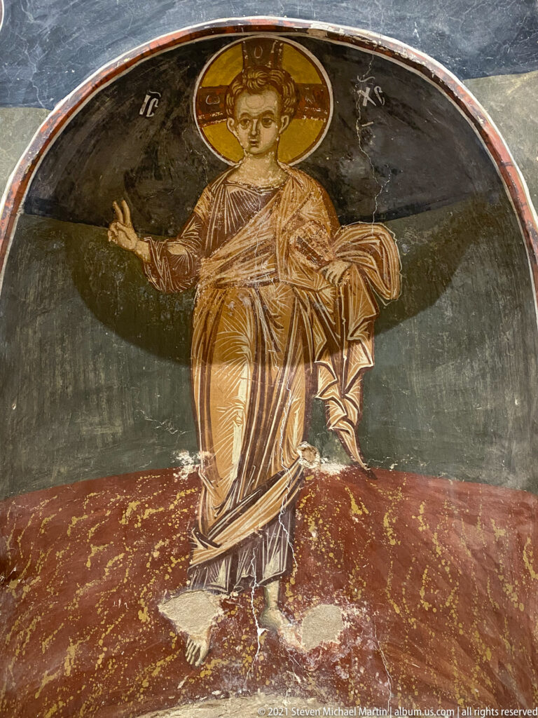 Front-facing haloed youthful saint seated barefoot beneath an arch with one hand raised.