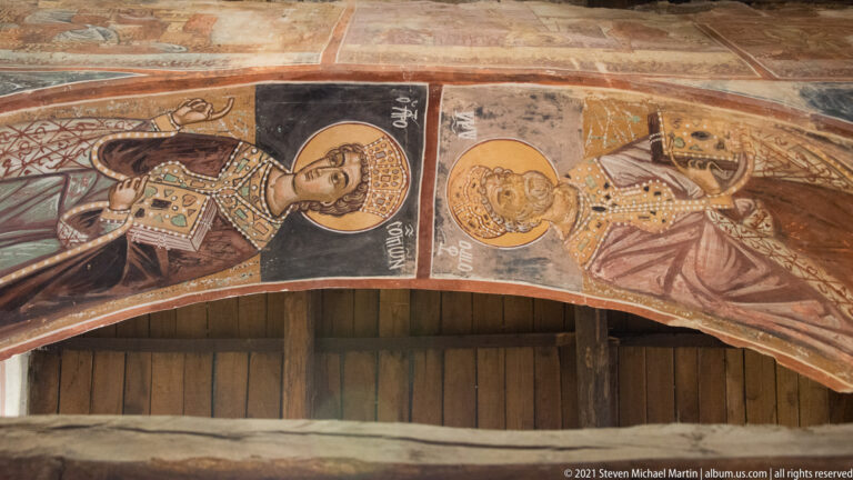 Two haloed crowned figures in richly patterned robes painted above a curved wooden arch.