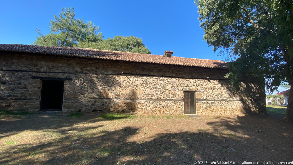 Long fieldstone church wall with red tile roof, open doorway, smaller wooden door, and large shade tree.