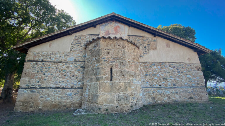 East end of the stone church with projecting polygonal apse, narrow opening, tiled gable, and faded mural above.