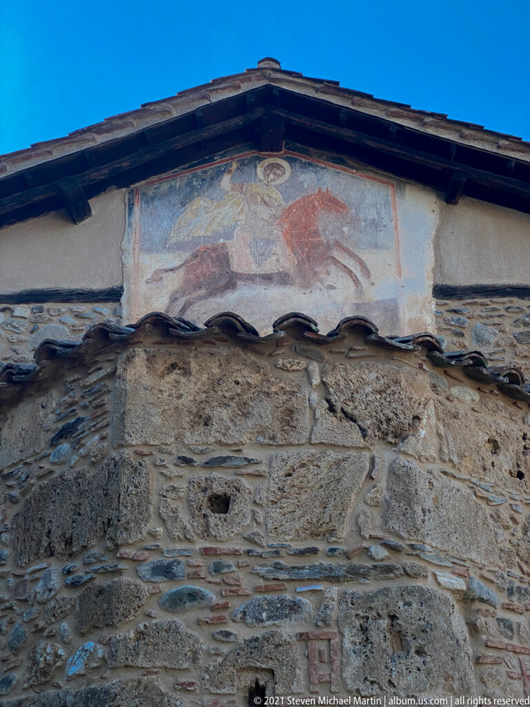 Faded gable fresco of a haloed mounted saint on a reddish horse, with a circular motif above.