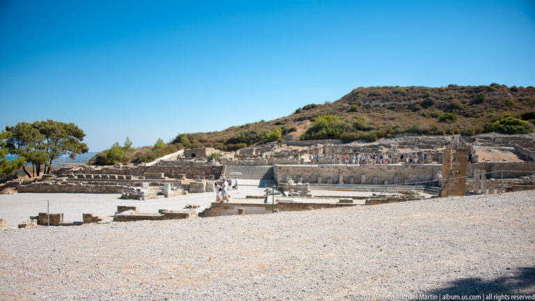 Wide view of Ancient Kamiros ruins with low stone walls, an open court, upright columns, and hillside beyond.