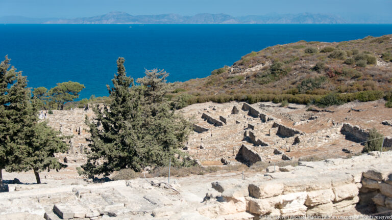 Ancient stone foundations and evergreen trees on a hillside overlooking the blue Aegean Sea and distant mountains.