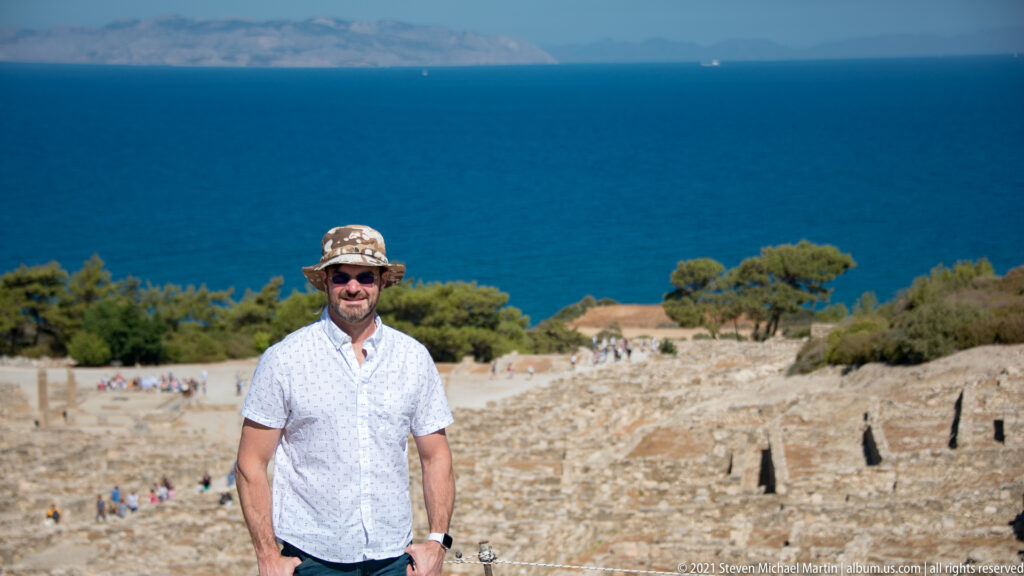 Man in a sun hat standing at Ancient Kamiros with excavated stone ruins and the blue Aegean Sea behind him.