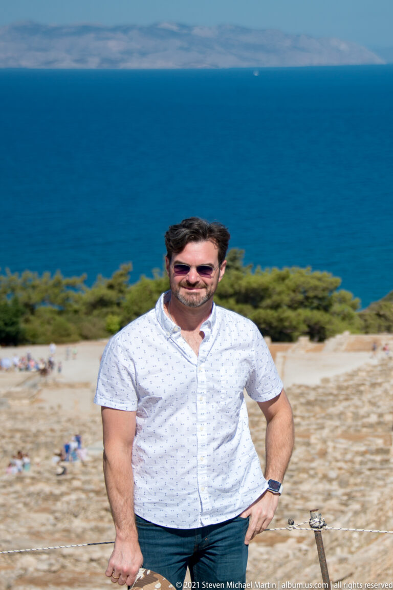 Close portrait of a man in a white shirt at Ancient Kamiros, with blurred ruins, trees, and blue sea behind.