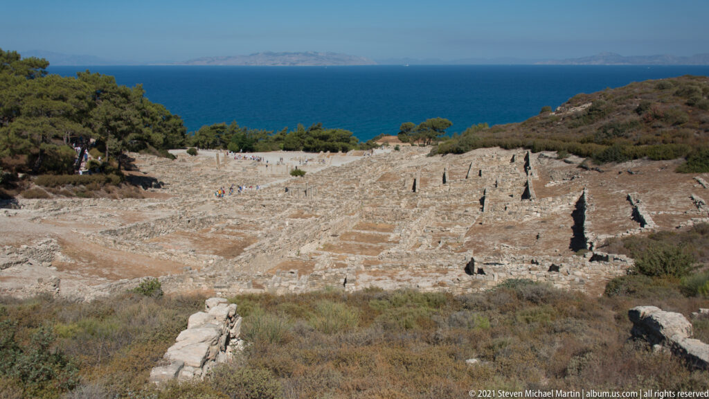 Wide hillside view of Ancient Kamiros residential foundations descending through scrub toward the blue Aegean Sea.