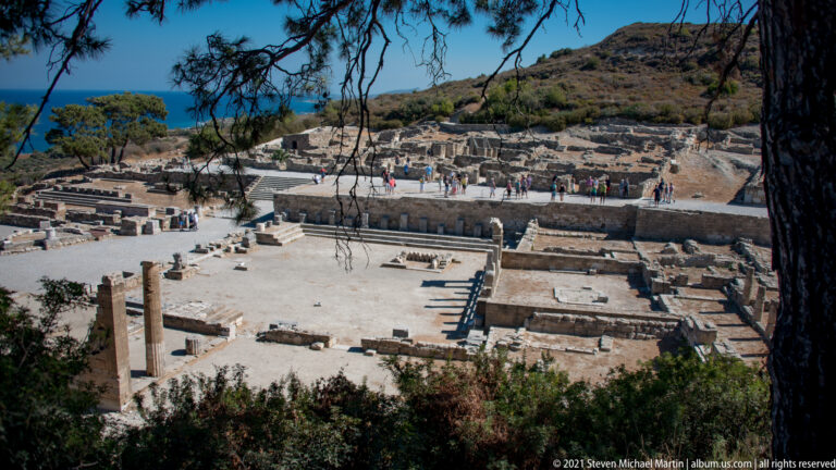 Elevated view over Ancient Kamiros showing rectangular stone foundations, passages, scattered columns, and visitors among the ruins.
