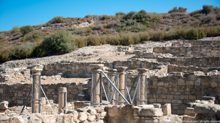 Standing ancient column shafts reinforced with modern metal braces amid low stone walls on the Ancient Kamiros hillside.
