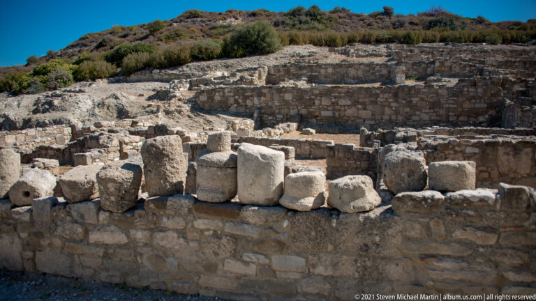 Loose carved and squared stone blocks arranged along a low wall with excavated ruins behind at Ancient Kamiros.