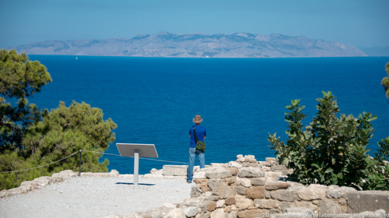 Visitor standing beside an interpretive stand at Ancient Kamiros, looking toward the blue Aegean Sea and distant mountains.
