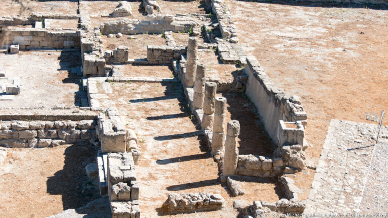Oblique view of several standing stone column shafts aligned beside a long wall and excavated rooms at Ancient Kamiros.