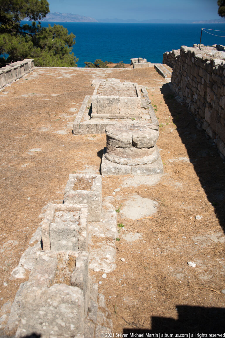 Long stone-paved passage or terrace through the ruins with the blue Aegean Sea beyond.