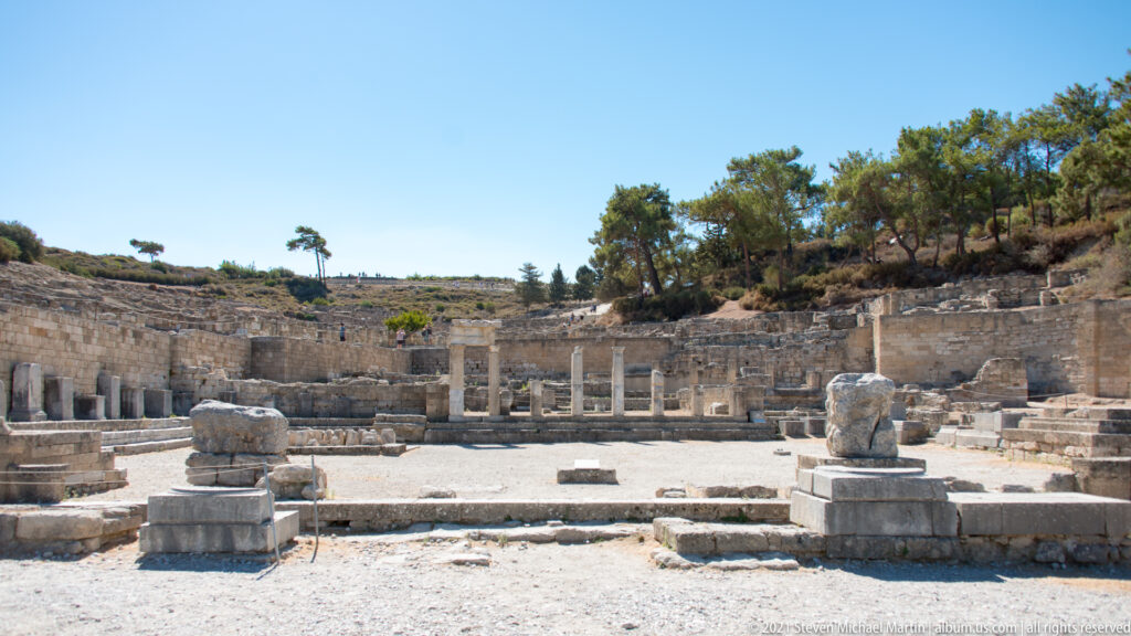 Broad archaeological precinct with standing columns, stone bases, low walls, and hillside terraces.