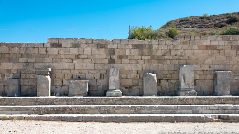 Row of stone pedestals and upright blocks on a terrace in front of a tall ashlar wall.