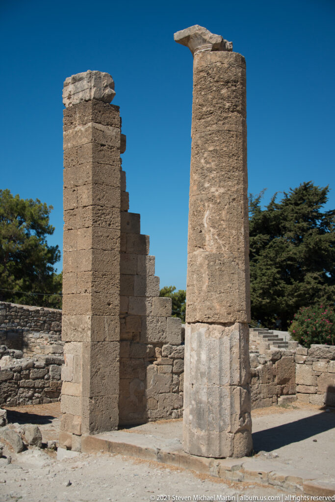 Two tall rectangular stone supports with projecting tops and pale restoration bands among ancient ruins.