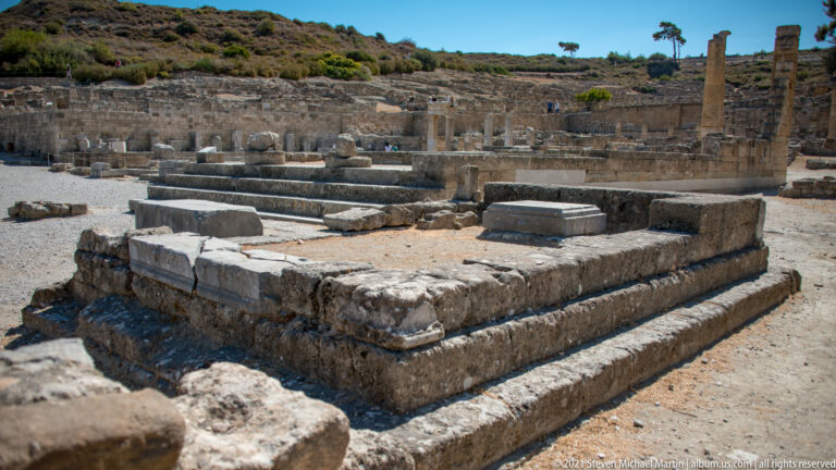 Large stepped rectangular stone platform in the foreground with columns and excavated foundations behind.