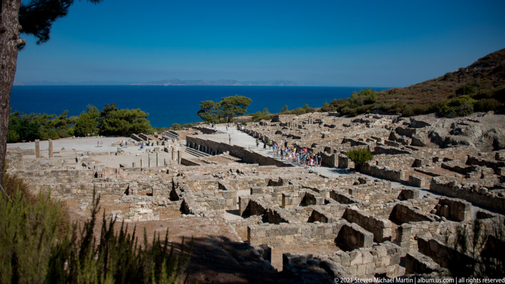 Panoramic hillside view of Ancient Kamiros ruins descending toward the blue Aegean Sea beneath a clear sky.