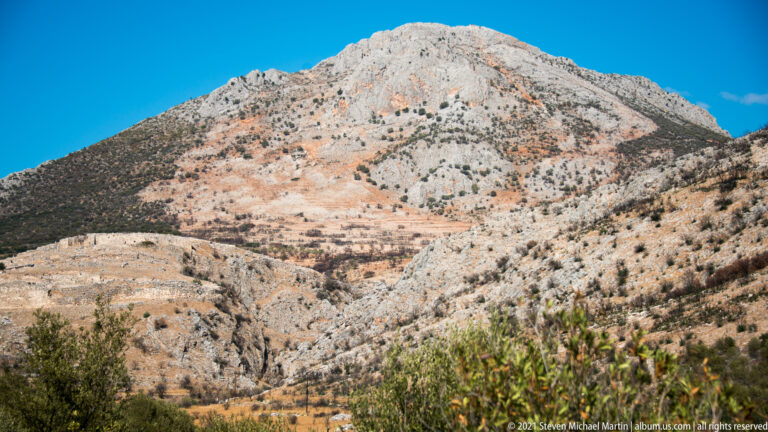 Rocky mountain rising above dry scrub and cultivated slopes near Ancient Mycenae under a clear blue sky.