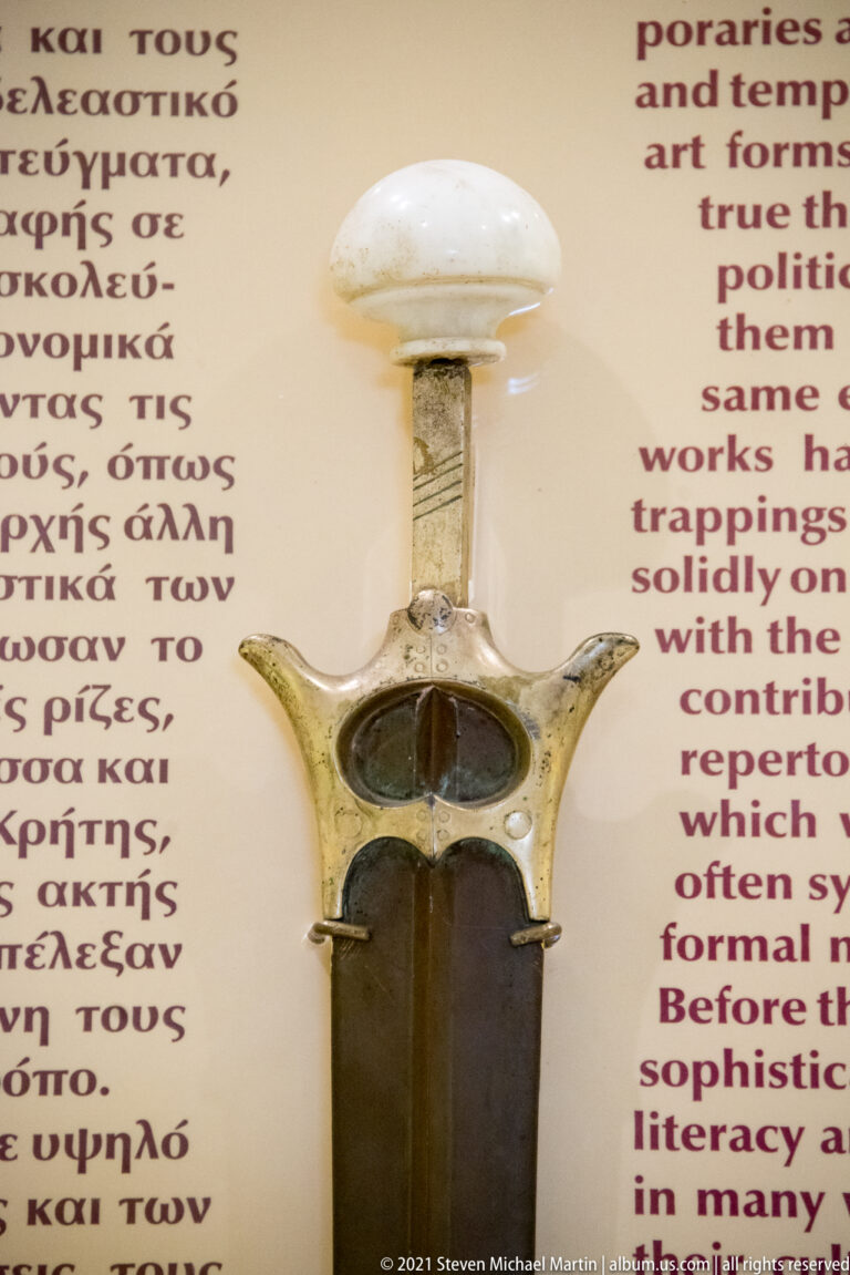 Small translucent ancient vessel with a long narrow neck, rounded rim, dark body, and two side projections.