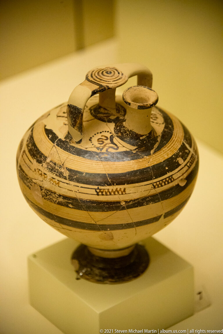 Rounded Mycenaean stirrup jar with bridge handle, offset spout, and dark concentric painted bands and spirals.