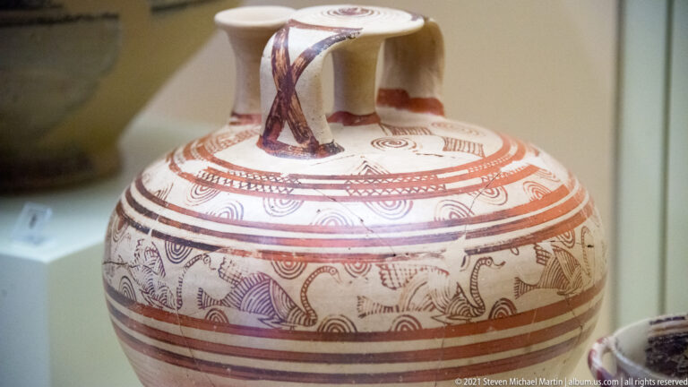 Cream ceramic jar with red-brown painted bands, spirals, and a multi-part bridge-like upper structure.
