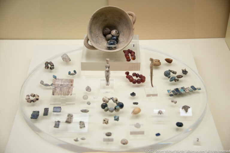 Museum case with beads, pendants, miniature figurines, plaques, and other small archaeological finds arranged on a round display.