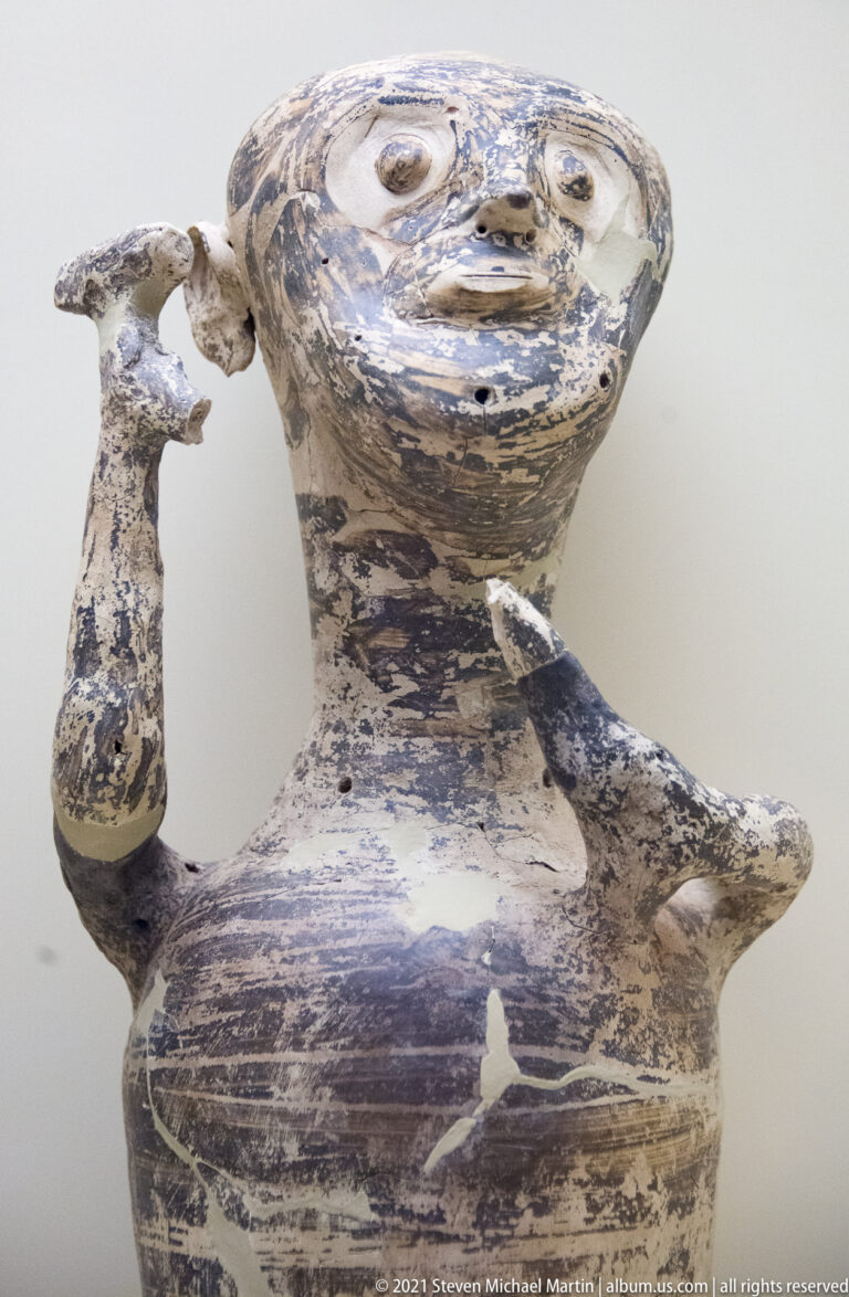 Anthropomorphic Mycenaean ceramic vessel with large eyes, projecting ears, and one arm raised beside the face.