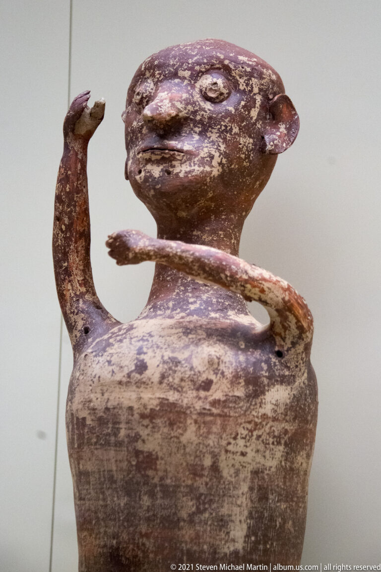 Mycenaean anthropomorphic ceramic vessel with a rounded head and two curved arms, one beside the face and one across the chest.