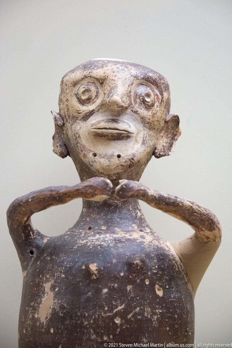 Dark-painted Mycenaean anthropomorphic vessel with round eyes and both arms curving across the upper torso.