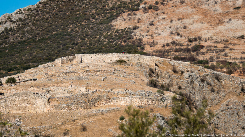 Low stone walls and foundations spread across a rocky hilltop terrace at Ancient Mycenae.