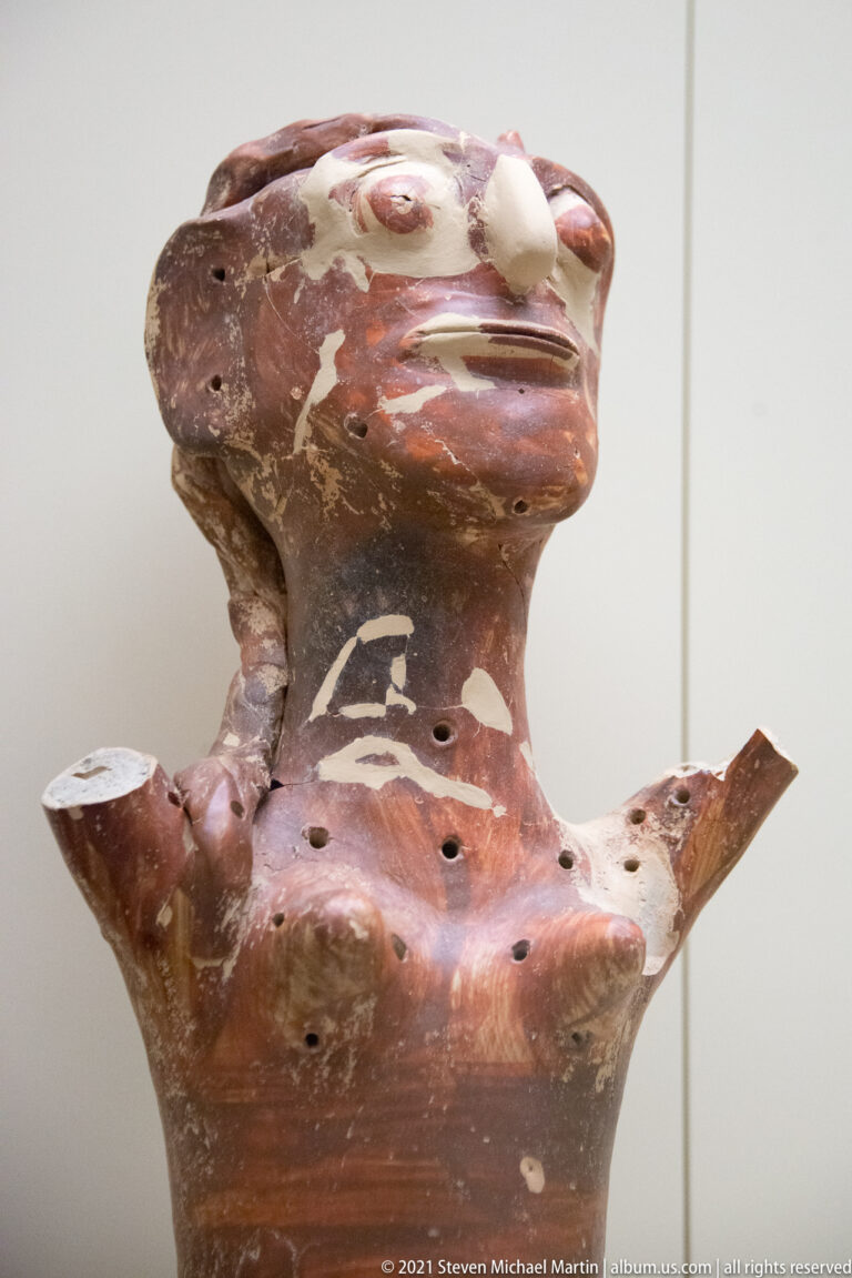 Red-painted Mycenaean anthropomorphic vessel with modeled face, projecting arms, white marks, and small torso perforations.