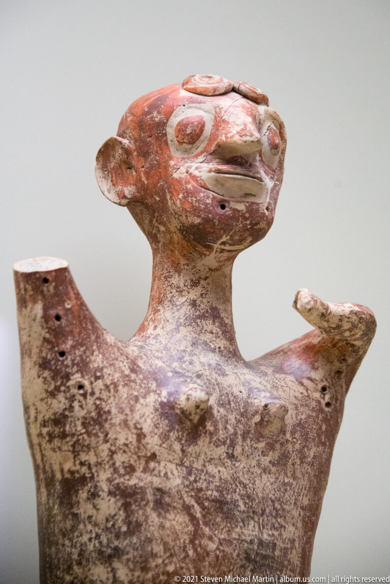 Mycenaean anthropomorphic ceramic vessel with long neck, modeled face, and both forearms raised outward.