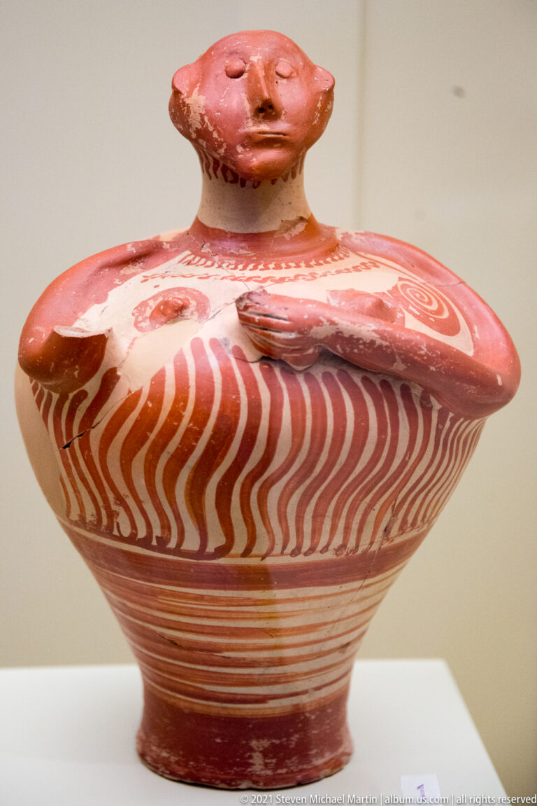 Mycenaean female ceramic figurine with small head, broad disk-like body, modeled breasts, and red painted wavy decoration.