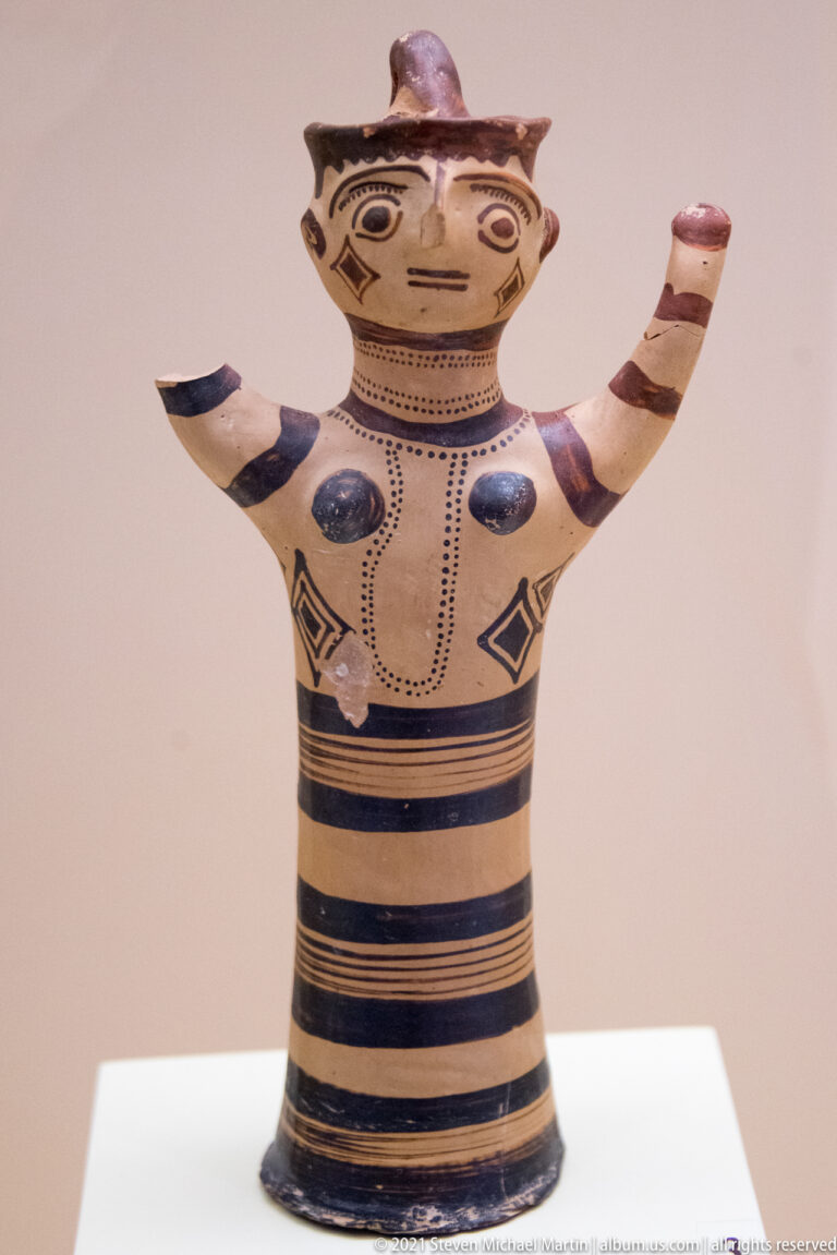 Standing Mycenaean female figurine with raised arms, modeled breasts, small head ornament, and blue-black painted bands and dots.