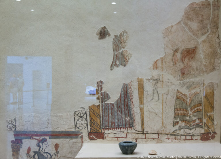 Fragmentary Mycenaean wall painting with surviving female garments, small figures, and scattered painted plaster patches on a pale wall.