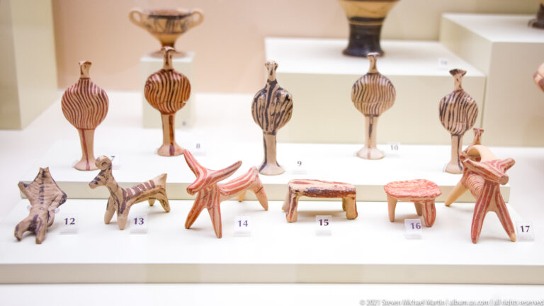 Rows of small painted terracotta figures and miniature stools or tables displayed together with numbered museum labels.