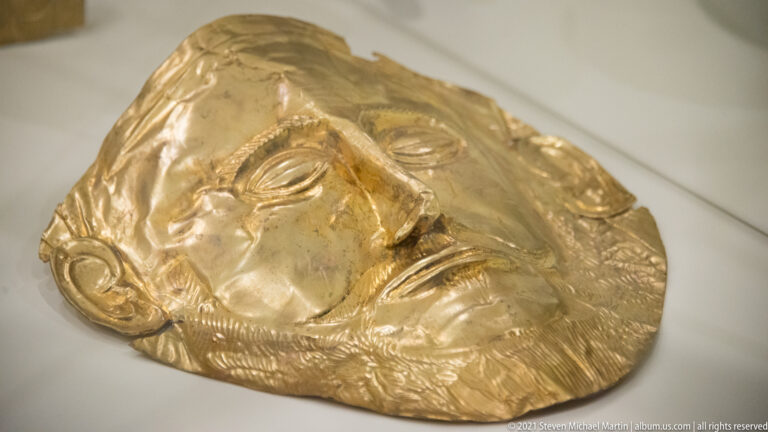Oblique view of a Mycenaean sheet-gold funerary mask with raised eyes, nose, lips, ears, and textured beard.