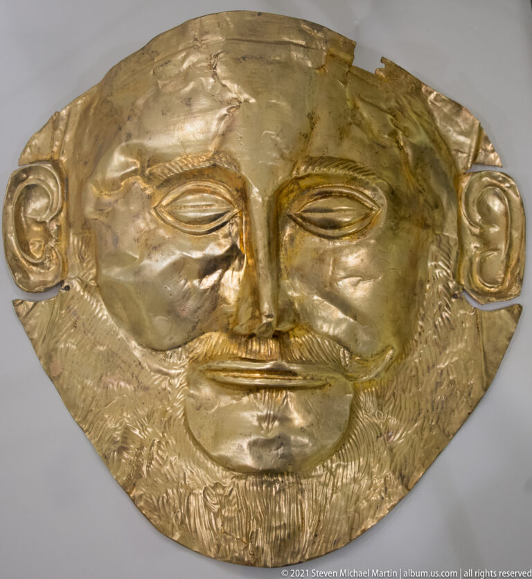 Frontal Mycenaean sheet-gold funerary mask with almond eyes, long nose, large ears, and textured moustache and beard.